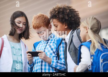Group of friends standing and looking at mobile phone of their classmate while he playing in it they standing outdoors after school lessons Stock Photo