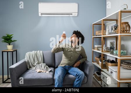 Young Happy Man Sitting On Couch Operating Air Conditioner With Remote Control At Home Stock Photo