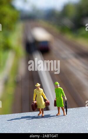 Two miniature female figurines standing on a plattform with luggage. In the blurry background are train tracks with some train leaving. Stock Photo