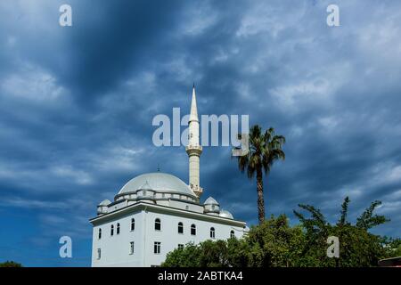Turkish landscape with white mosque and minaret on a dark blue cloudy sky background. On the right side are tops of the palm trees. Stock Photo