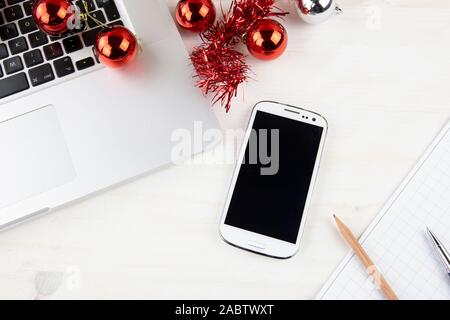 Computer job at Christmas holidays concept: close up top view of an aluminum laptop open, smartphone, red wreath decoration, red and silver baubles, p Stock Photo