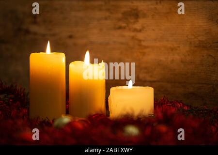 The warmth of the Christmas concept: three candles lit on a light wooden table and a rustic setting with pine branches, red decoration and gold and re Stock Photo