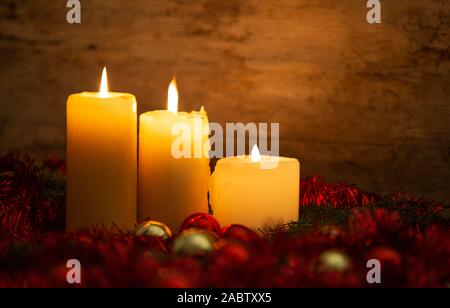 The warmth of the Christmas concept: three candles lit on a light wooden table and a rustic setting with pine branches, red decoration and gold and re Stock Photo