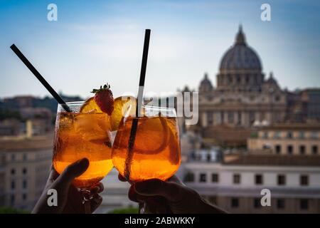 A couple is holding glasses of Aperol in the bar inside the Castle Sant'Angelo. Glasses of Aperol and St Peter's basilica at the background. Stock Photo