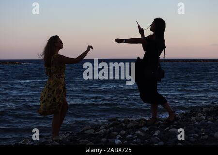 Two young girls from East Europa on beach silhouetted taking pictures with their  smartphones at sunset