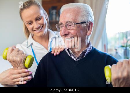 Female Physiotherapist Helping Senior Man To Lift Hand Weights At Home Stock Photo