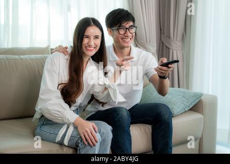 Asian young loving couple sitting and watching TV and smile when watching fun program in TV in a comfortable couch in the livingroom at home. Family l Stock Photo