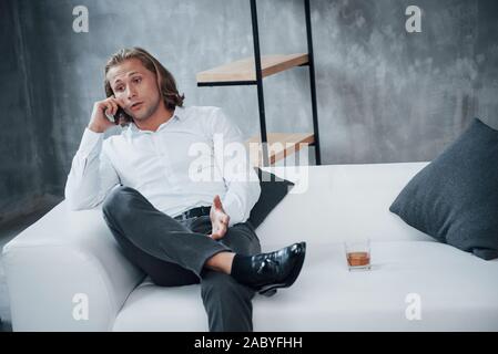 Portrait of young businessman with long hair sitting on couch and talking on mobile phone with whiskey on right side Stock Photo
