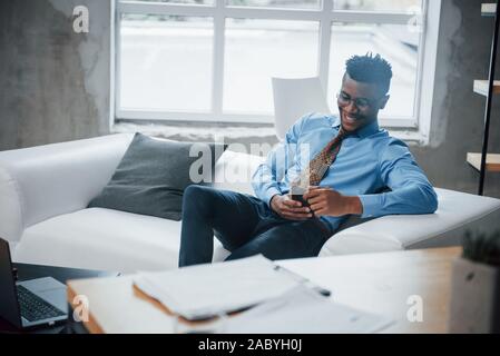 Smiling afroamerican guy sitting on the couch and looking at his smartphone Stock Photo