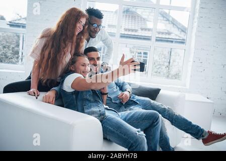 Happy faces. Cheerful young friends taking selfies on sofa and white interior Stock Photo