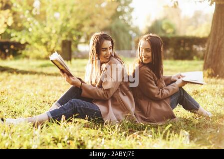 Smiling brunette twin girls sitting back to back on the grass and looking at each other, legs slightly bent in knees, with brown books in hands Stock Photo
