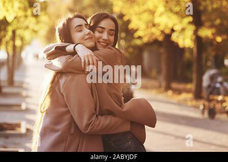 Side profile of young pretty smiling brunette twin girls hugging and having fun in casual coat standing close to each other at autumn sunny park alley Stock Photo