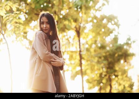 Young happy brunette girl standing and slightly hugging herself with hands in front, wearing casual coat with autumn yellow trees on the light blurry Stock Photo