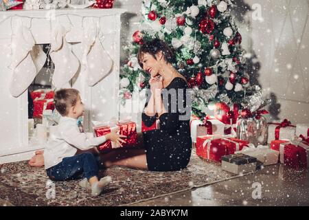 https://l450v.alamy.com/450v/2abyk7m/son-gives-a-christmas-gift-to-mom-mom-pleasantly-surprised-and-cute-looks-at-the-child-isolated-on-a-new-year-tree-background-with-gifts-a-fireplac-2abyk7m.jpg