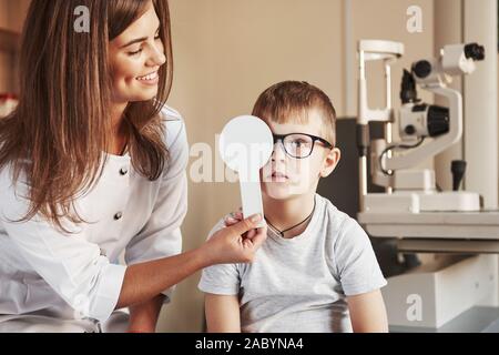 Concentrating boy. Female doctor covers kid eye with medical tool for checking visual acuity