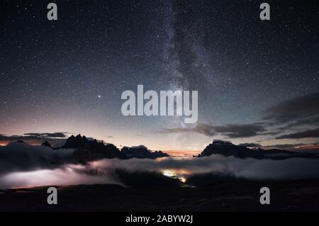 You can clear see the milky way. Something is shining in the middle of the forest with stars in the sky. The mountains are surrounded by dense fog Stock Photo