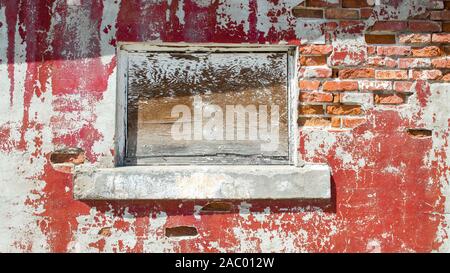boarded up window in a crumbling wall of an abandoned building with red peeling paint and exposed bricks on a sunny day Stock Photo