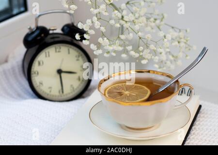 Gypsophila bouquet on the window in a glass vase at dusk. Cup of tea with lemon. The clock shows half past three. Stock Photo