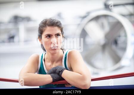 A badass female boxer is resting on ring ropes with her wraps on in a boxing gym, white walls, black and red mats, indoors. Stock Photo