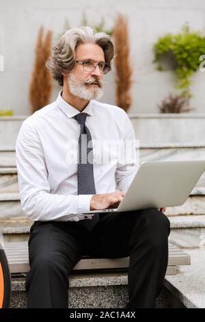 Photo of handsome focused businessman in eyeglasses typing on laptop while sitting on bench outdoors Stock Photo