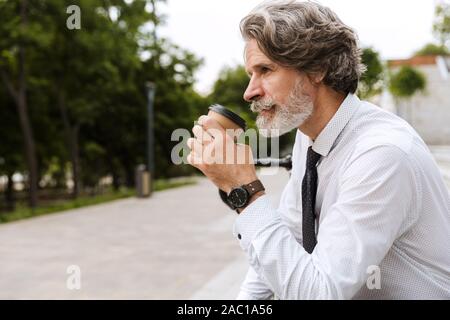 Photo of mature bearded businessman in formal clothes drinking coffee takeaway while sitting outdoors Stock Photo