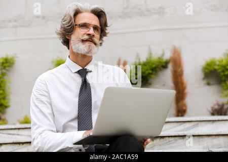 Photo of handsome gray-haired businessman in eyeglasses typing on laptop while sitting outdoors Stock Photo