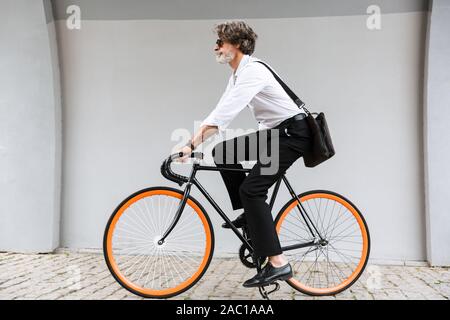 Photo of modern old businessman in sunglasses riding bicycle over gray wall on city street Stock Photo