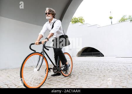 Photo of confident elderly businessman in sunglasses and white shirt riding bicycle on city street Stock Photo