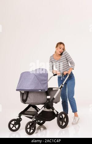Full length of a beautiful young woman standing with a baby pram isolated over white background, talking on mobile phone Stock Photo