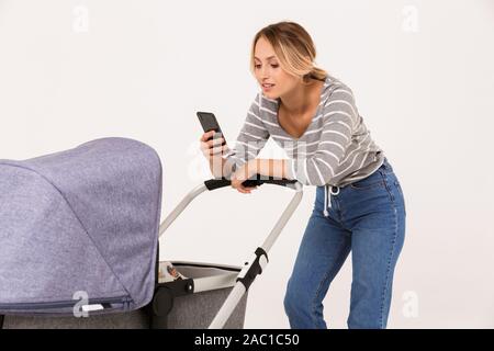 Beautiful young woman standing with a baby pram isolated over white background, using mobile phone Stock Photo