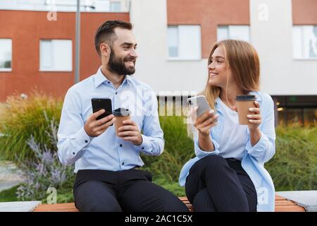 Image of a young happy colleagues man and woman businesspeople sit outside at the street using mobile phones talking with each other. Stock Photo