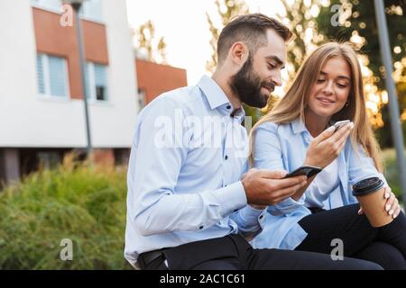 Photo of a young cheery man and woman businesspeople sit outside at the street using mobile phones talking with each other. Stock Photo