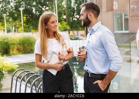Image of a happy young business couple colleagues with cup of coffee and notebook outdoors near business center talking with each other. Stock Photo