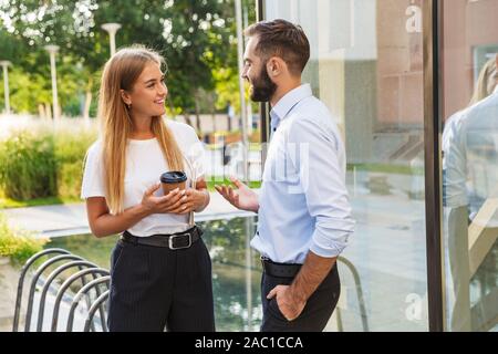 Image of a young positive smiling man and woman businesspeople outside at the street near business center holding cup of coffee talking with each othe Stock Photo