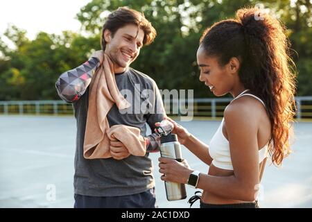 Image of a handsome positive smiling young strong sports man holding towel talking with fitness woman holding water outdoors. Stock Photo
