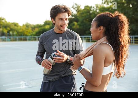 Image of a handsome positive smiling young strong sports man holding towel talking with fitness woman holding water outdoors. Stock Photo