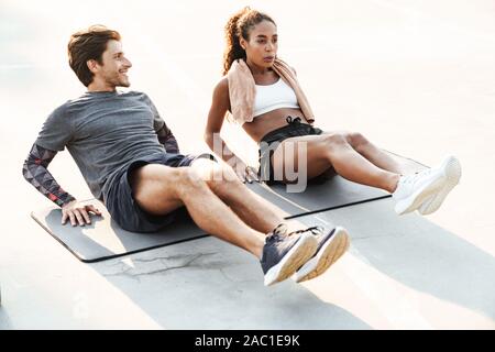 Photo of multinational young couple in sportswear doing exercise on mat while working out at playground outdoors Stock Photo