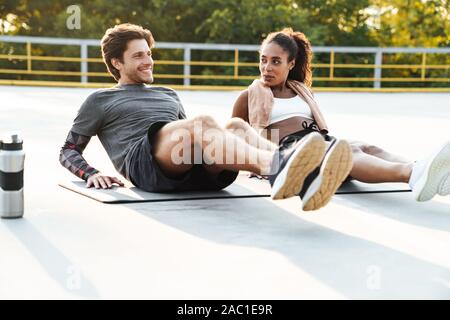 Photo of young cheerful couple in sportswear doing exercise on mat while working out at playground outdoors Stock Photo