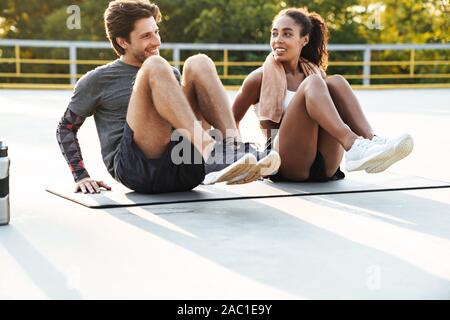 Photo of multinational cheerful couple in sportswear doing exercise on mat while working out at playground outdoors Stock Photo