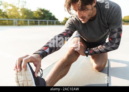 Photo of athletic caucasian man in sportswear stretching his leg on mat while working out at playground outdoors Stock Photo