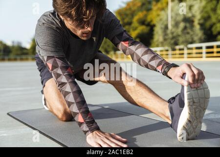 Photo of athletic unshaven man in sportswear stretching his leg on mat while working out at playground outdoors Stock Photo