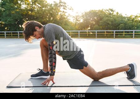 Photo of athletic young man in sportswear stretching his legs on mat while working out at playground outdoors Stock Photo