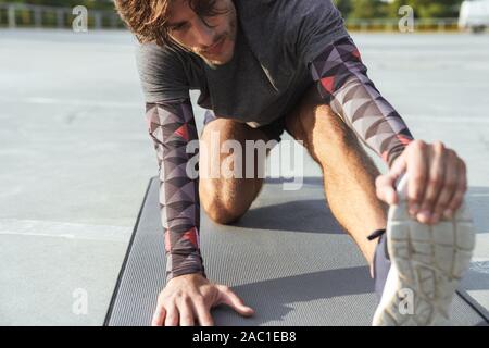 Photo of strong caucasian man in sportswear stretching his leg on mat while working out at playground outdoors Stock Photo