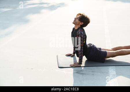 Photo of young focused man in sportswear doing yoga exercises on mat while working out at playground outdoors Stock Photo
