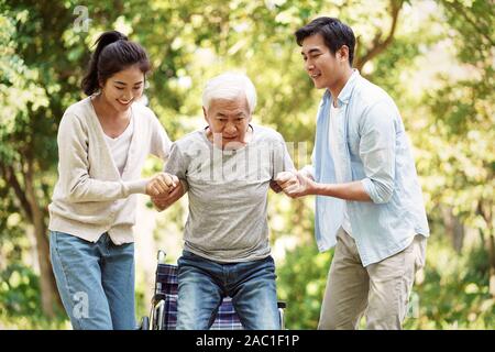 young asian man and woman helping wheelchair bound senior man stand up and walk Stock Photo