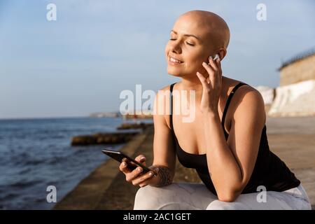 Image of happy bald woman in sportswear using cellphone and earphone while sitting near seaside in sunny morning Stock Photo