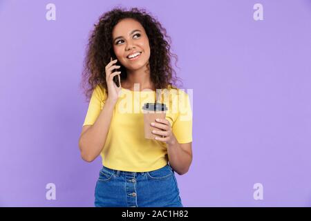Image of pretty african american woman with curly hair holding coffee cup and talking on smartphone isolated over violet background Stock Photo