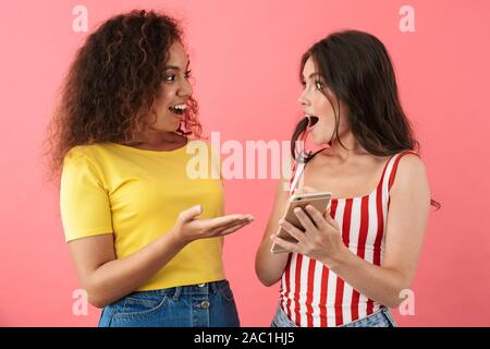Image of surprised multinational girls smiling and holding cellphones together isolated over pink background Stock Photo