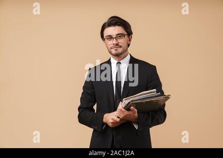 Portrait of a handsome smiling confident young businessman wearing suit standing isolated over beige background, holding stack of documents Stock Photo
