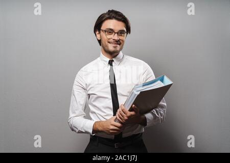 Portrait of a handsome young businessman wearing suit standing isolated over gray background, holding binder, lookimng at camera Stock Photo